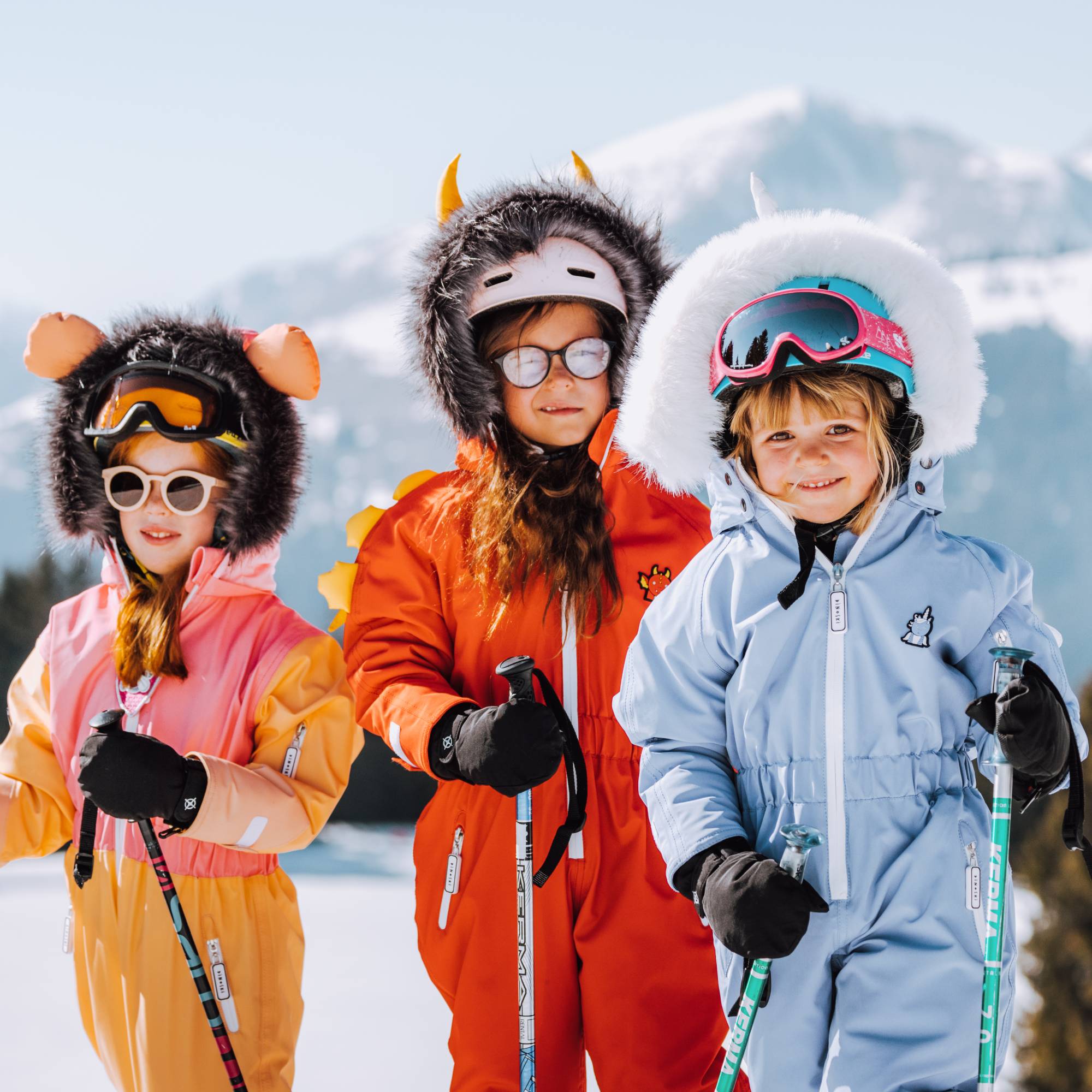Drei Kinder tragen bunte Schneeanzüge von LibertyKids – bereit für den Winterurlaub, vor verschneiter Alpenkulisse.