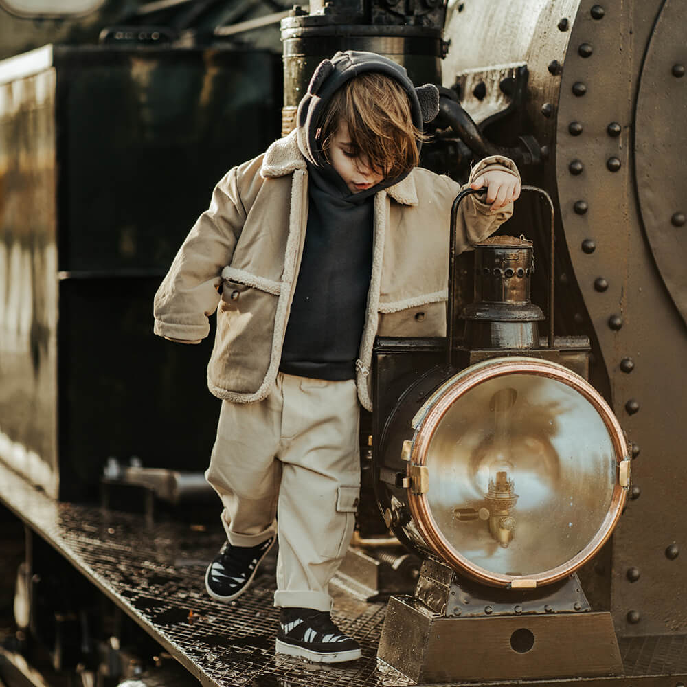 Kind in beiger Donsje-Kleidung balanciert auf einer alten Lokomotive – Black Friday Kinder-Mode mit bis zu 25 % Rabatt bei LibertyKids