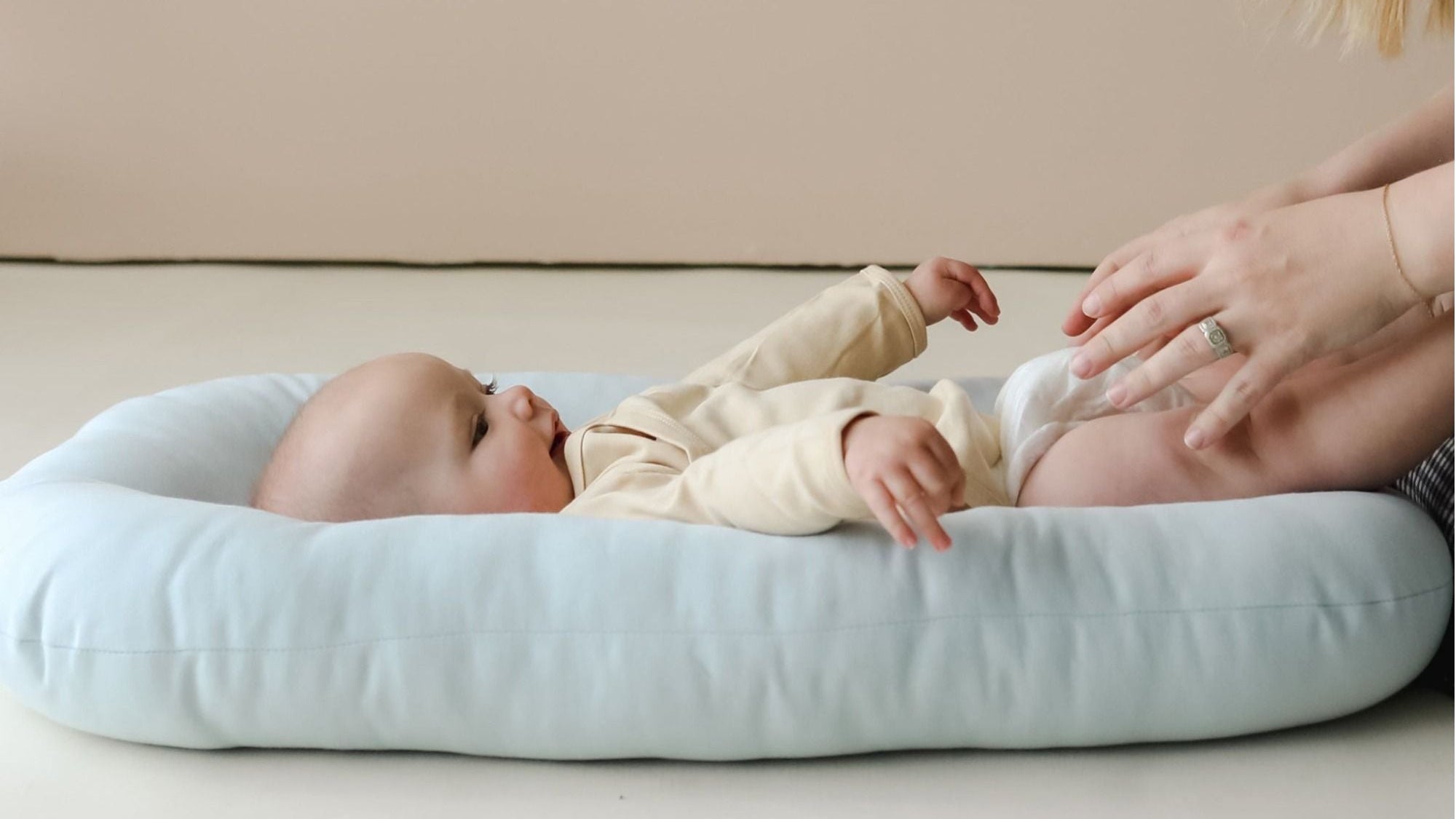 Ein Baby liegt entspannt in einem hellbeigen Snuggle Me Organic Babynest Lounger auf einem hellen Hintergrund.