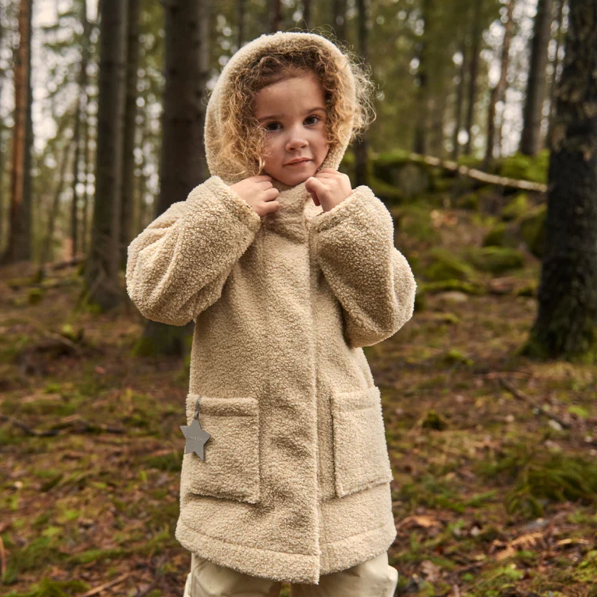 Ein Kind steht im Wald und trägt einen warmen, beigefarbenen Teddyfell-Wintermantel mit Kapuze von Mini A Ture – ideal für die kalte Jahreszeit.