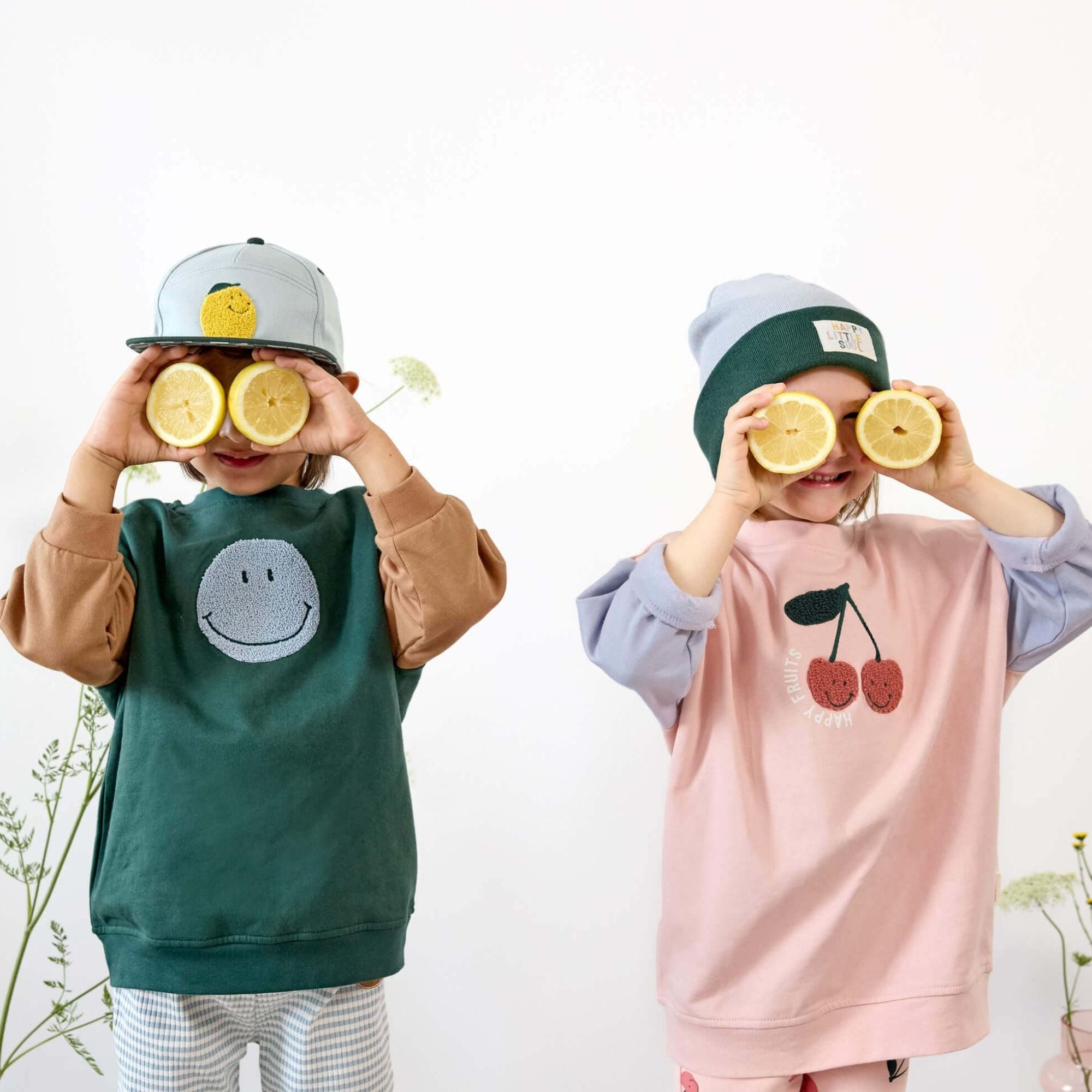 Zwei Kinder mit bunten Lässig-Pullovern und Mützen halten Zitronen vor die Augen – fröhlich, verspielt, kindgerecht.