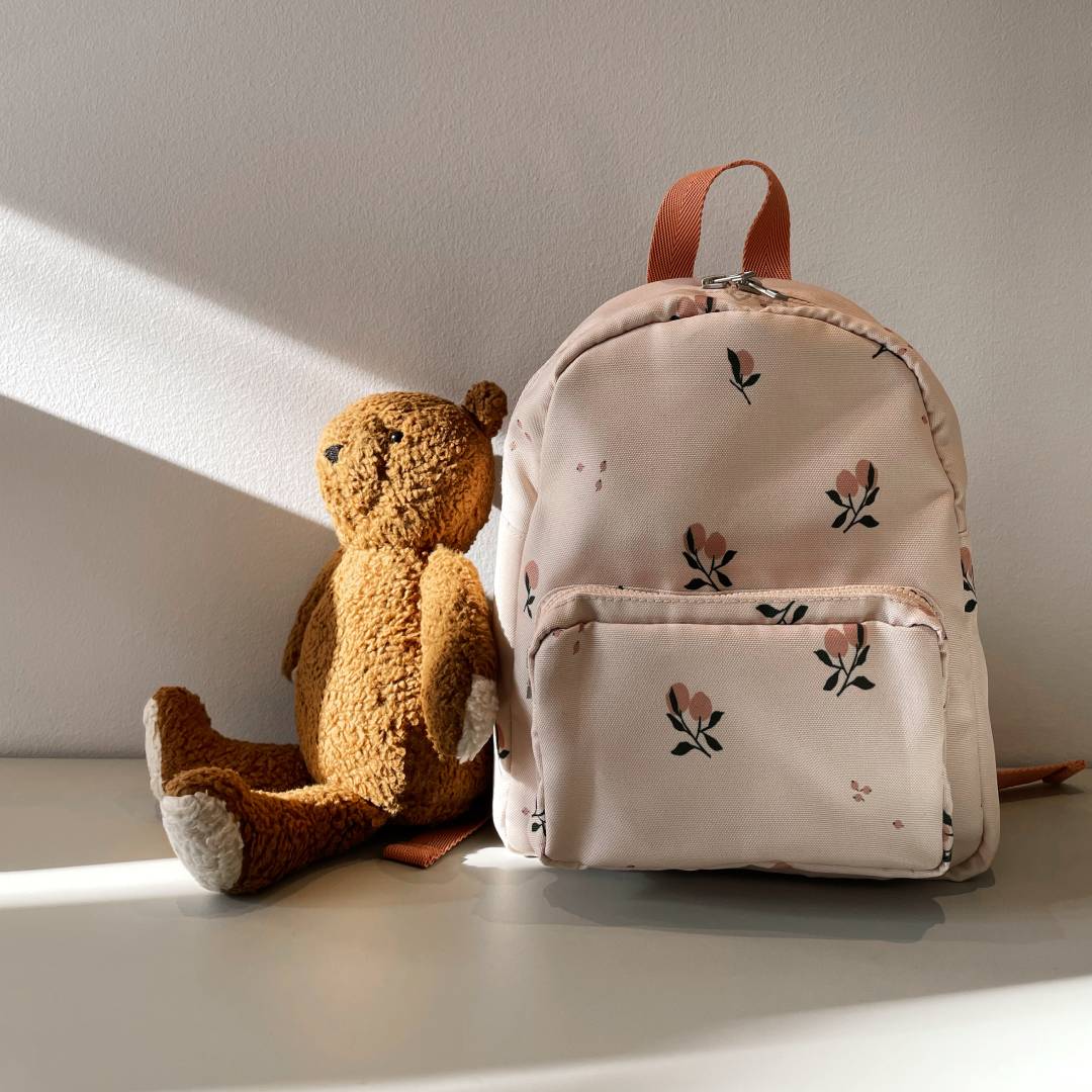Kinder-Rucksack mit Blumenmuster steht neben einem Stoffteddy im Sonnenlicht auf einer weißen Oberfläche.
