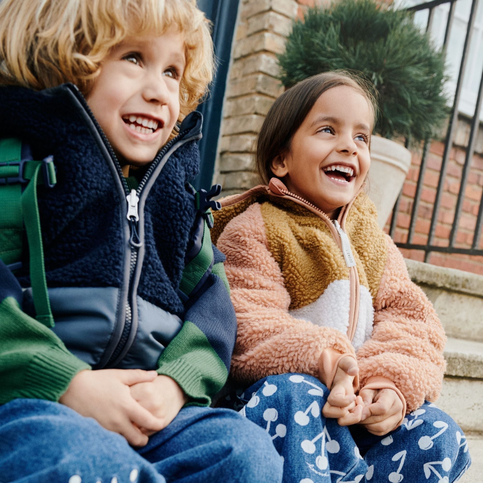Zwei fröhliche Kinder in farbenfroher Winterkleidung von LIEWOOD sitzen auf einer Treppe und lachen.