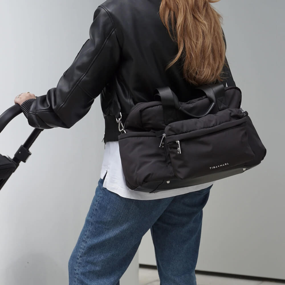 Frau mit stylischer schwarzer Wickeltasche von TIBA + MARL am Kinderwagen – modernes Eltern-Accessoire im minimalistischen Design