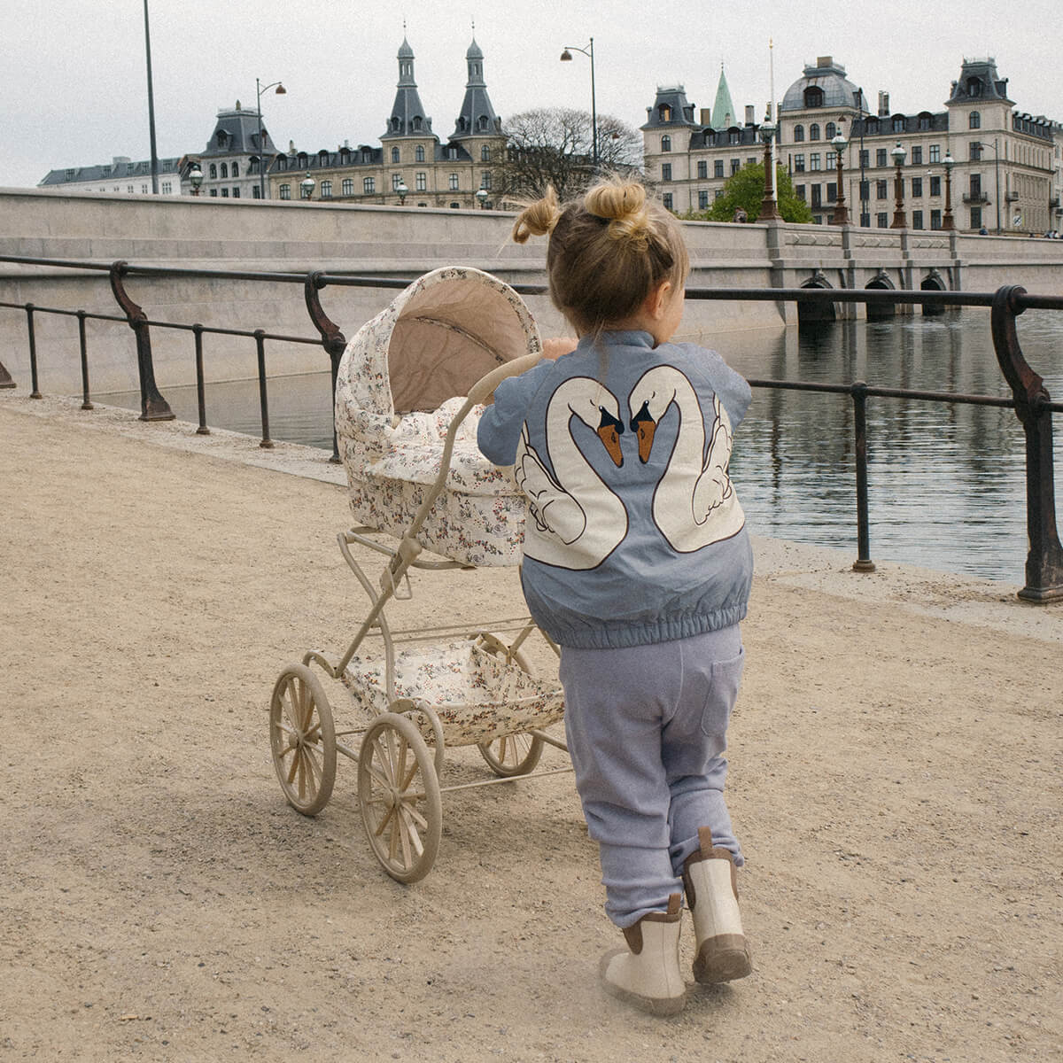 Kleines Kind schiebt einen Puppenwagen am Flussufer entlang, trägt eine blaue Jacke mit zwei weißen Schwänen auf dem Rücken.