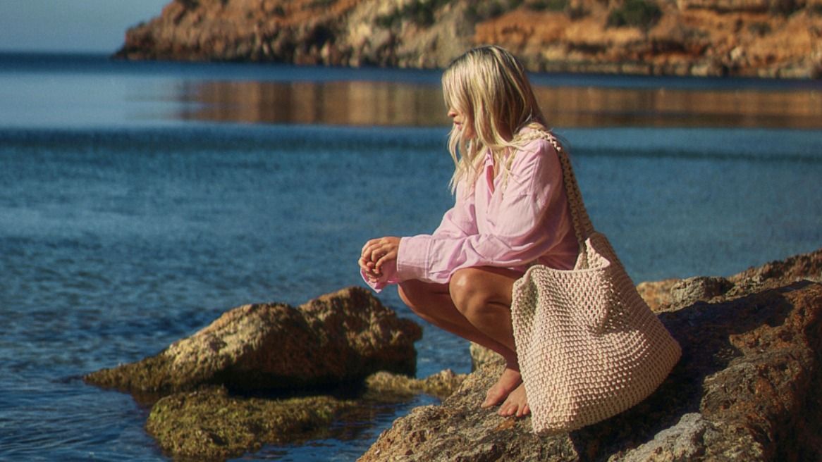 Frau sitzt barfuß auf Felsen am Meer mit einer natürlichen Macramé-Tasche von LibertyKids – ideal für Strandtage