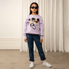 Mädchen mit Sonnenbrille trägt einen lilafarbenen Pullover mit Bärchen-Print, kombiniert mit lässiger Jeans – steht vor weißem Vorhang