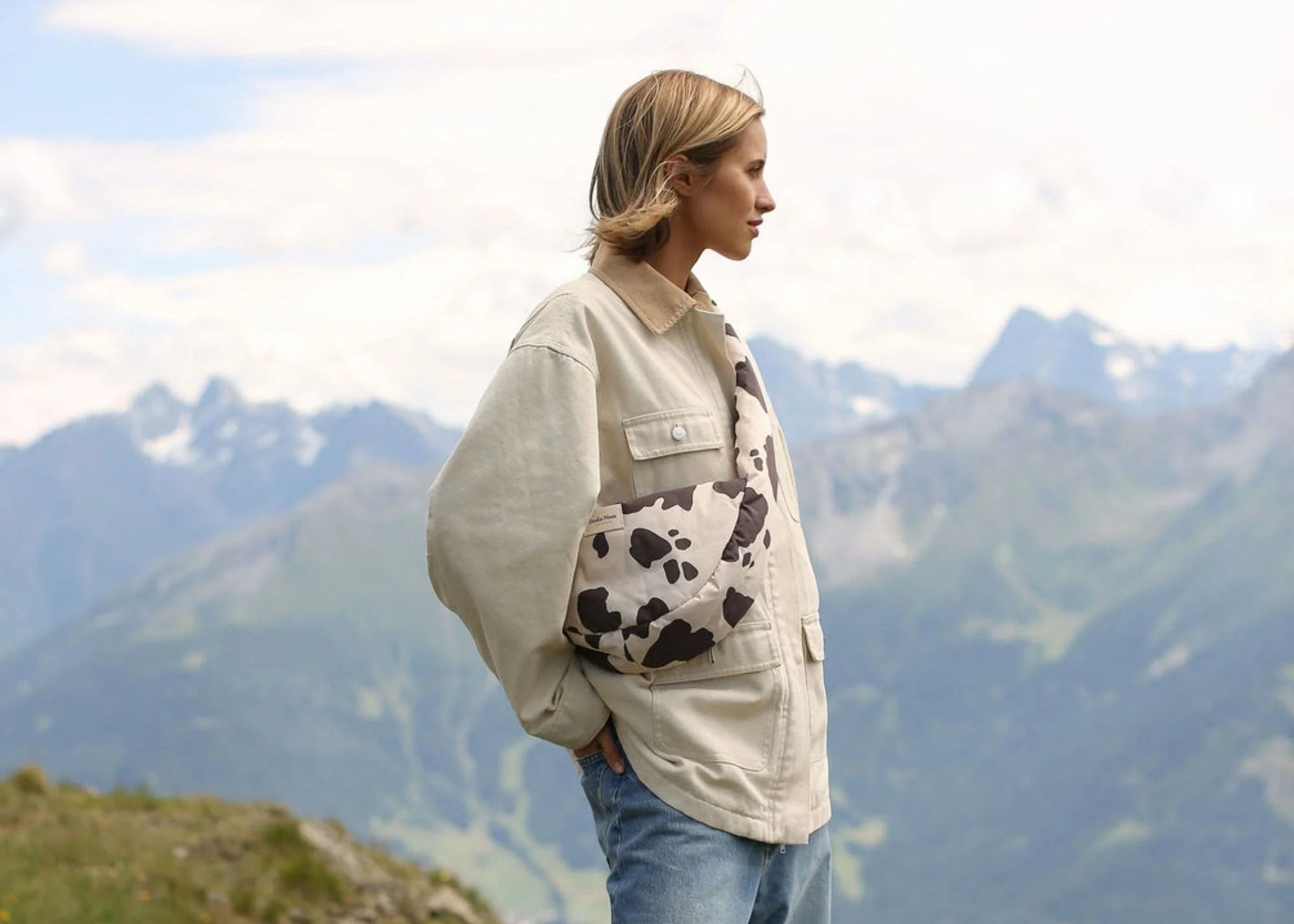 Eine Frau in den Bergen trägt die Studio Noos Adult Crossbody Bag im Cow Print Design lässig über der Schulter.