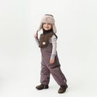 Kind mit braunStrickschal, flauschiger Fellmütze und Latzhose – warmer Winter-Look mit Stil