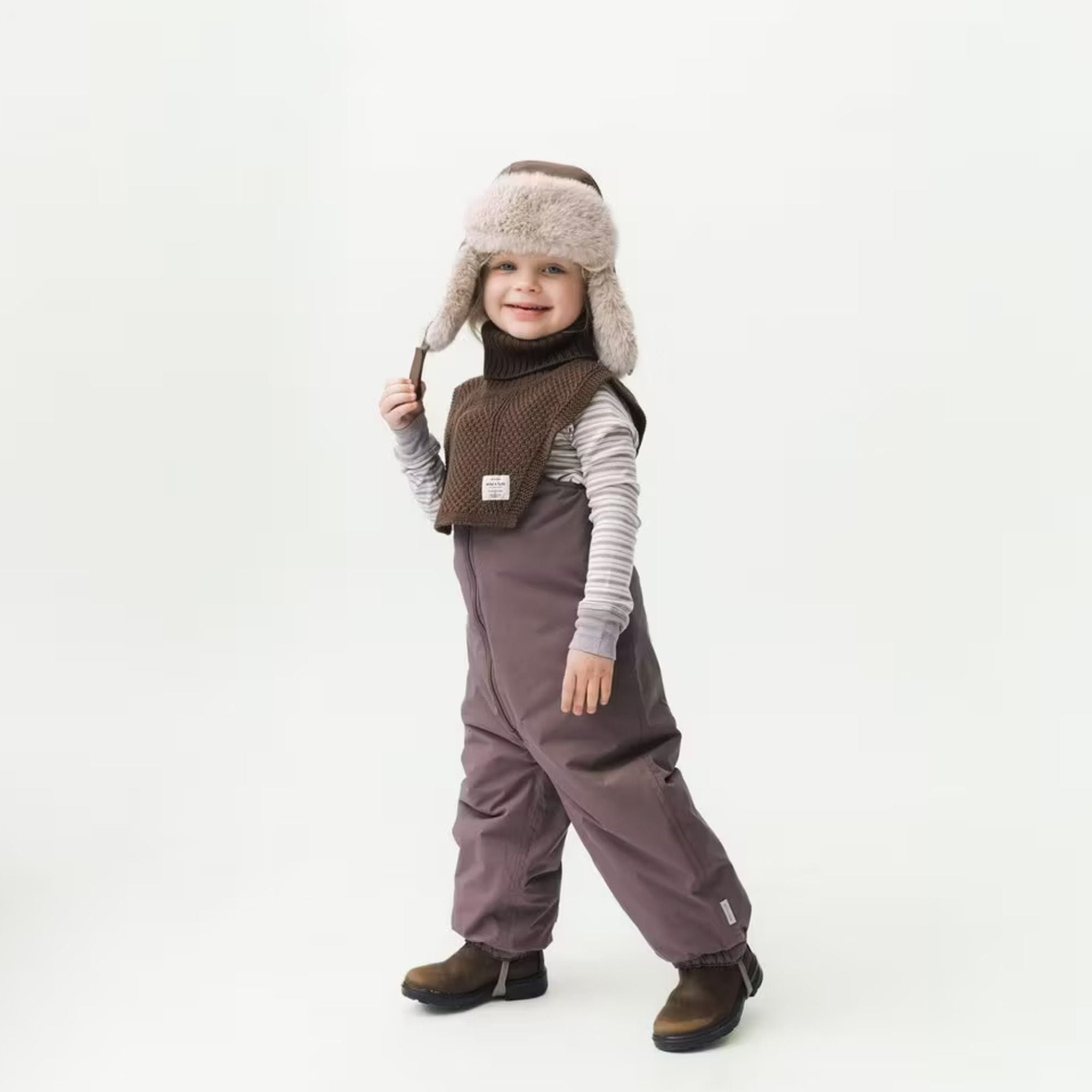 Kind mit braunStrickschal, flauschiger Fellmütze und Latzhose – warmer Winter-Look mit Stil